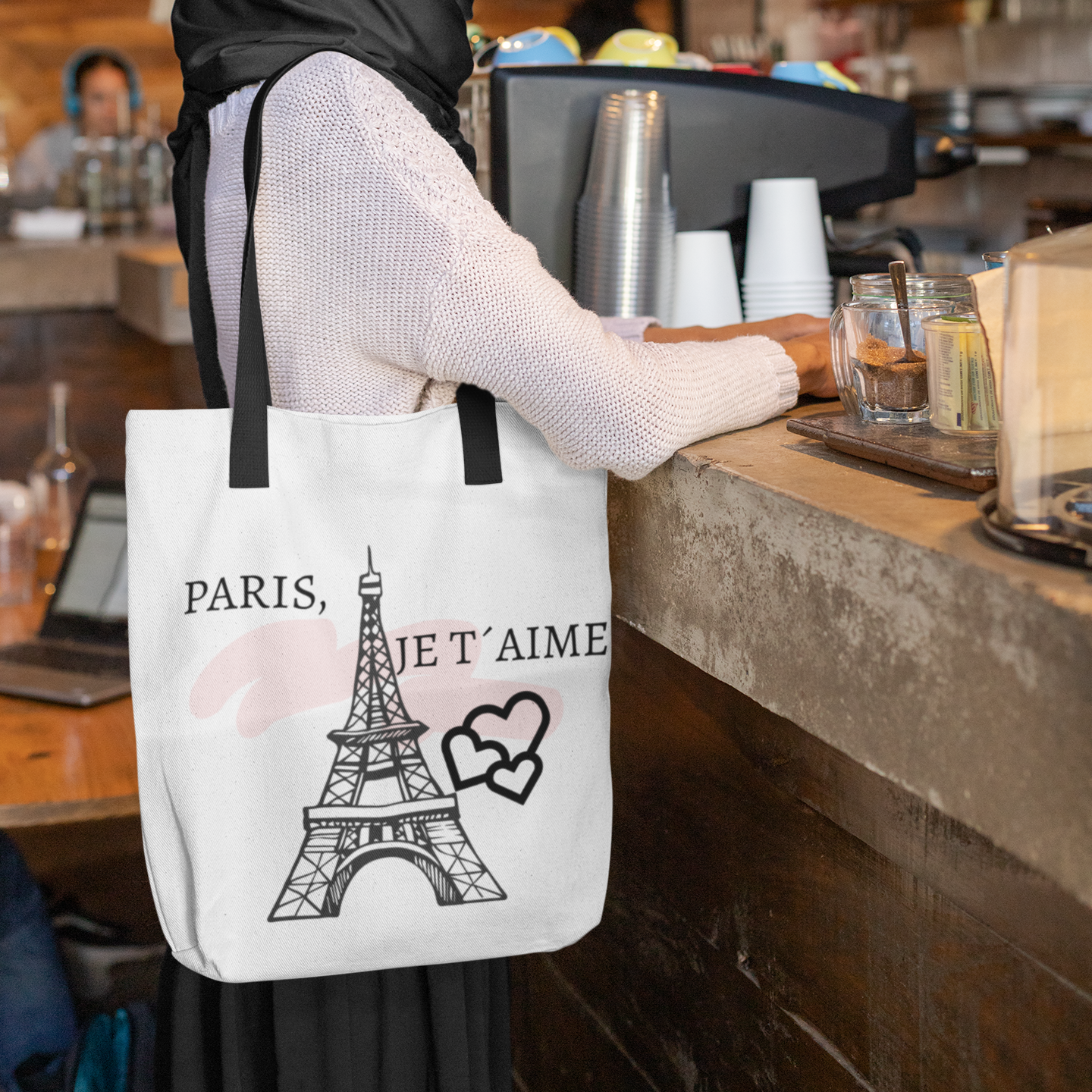 Woman in a cafe wearing a tote bag with the Eiffel Tower and 'Paris, je t'aime' text.